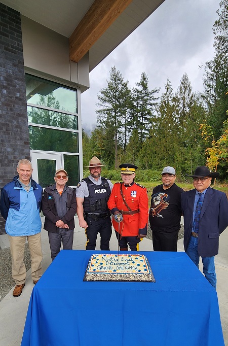 Michael Lott, le maire du village de Gold River; Martin Davis, celui du village de Tahsis; le sergent Greg Young, chef du Détachement de la baie Nootka; les chefs Nathan George et Jerry Jack de la Première Nation Mowachaht/Muchalaht; et le sous-commissaire Dwayne McDonald, commandant divisionnaire de la GRC en C.-B., debout au milieu en train de couper le gâteau cérémoniel.