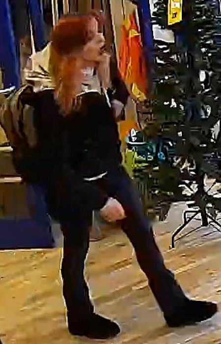 Une femme aux cheveux roux, portant un manteau et un pantalon foncés et un chandail blanc, marche près d’un arbre de Noël dans un magasin de détail.